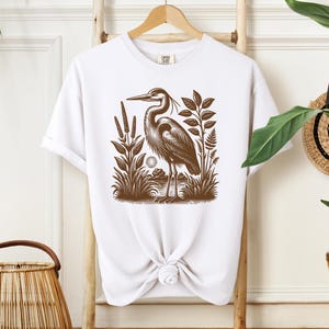 May include: White t-shirt with a brown illustration of a heron standing in a marsh with reeds and plants.