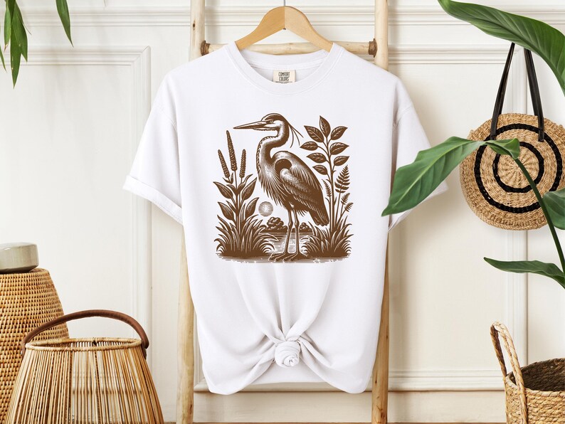 May include: White t-shirt with a brown illustration of a heron standing in a marsh with reeds and plants.