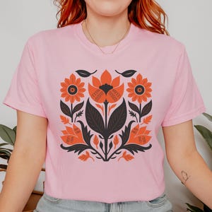 May include: A light pink t-shirt featuring a symmetrical floral design in orange, black, and white. The design includes stylized flowers, leaves, and stems, creating a bold, graphic pattern. The shirt is a crew neck style.