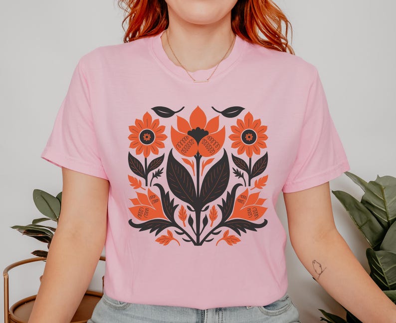 May include: A light pink t-shirt featuring a symmetrical floral design in orange, black, and white. The design includes stylized flowers, leaves, and stems, creating a bold, graphic pattern. The shirt is a crew neck style.