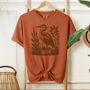 May include: A rust-colored t-shirt with a vintage illustration of a heron standing in a marsh. The heron is brown and the marsh is green.