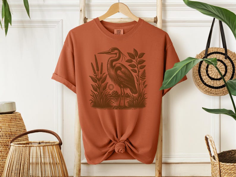 May include: A rust-colored t-shirt with a vintage illustration of a heron standing in a marsh. The heron is brown and the marsh is green.