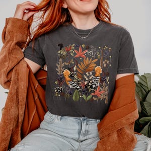 May include: A dark gray t-shirt with a colorful illustration of two hoopoes surrounded by flowers and leaves. The illustration is in a square shape and is centered on the shirt.