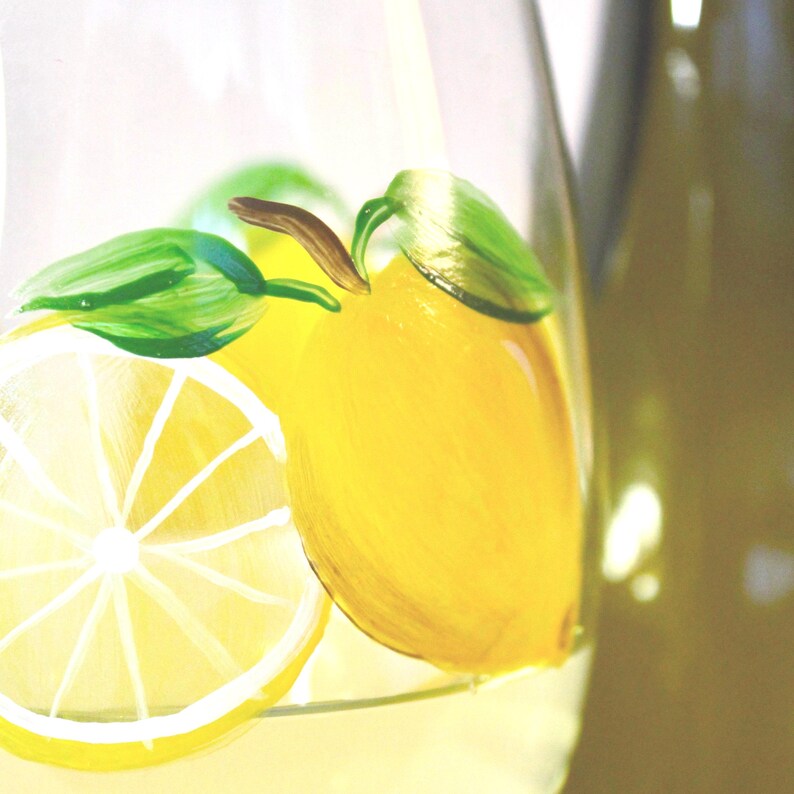 May include: A clear glass pitcher with a hand-painted lemon design. The design features a whole yellow lemon, a lemon slice, and green leaves. The pitcher is partially filled with a yellow liquid, suggesting lemonade or a similar beverage.
