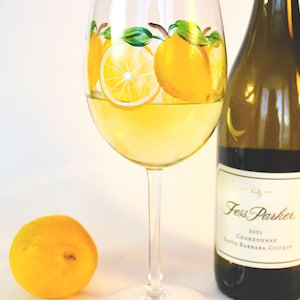 May include: A wine glass filled with yellow liquid, decorated with painted lemons and green leaves. A bottle of Fess Parker Chardonnay is next to the glass, and a whole lemon sits in front. The glass has a lemon slice painted on the base.