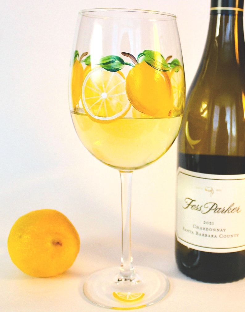 May include: A wine glass filled with yellow liquid, decorated with painted lemons and green leaves. A bottle of Fess Parker Chardonnay is next to the glass, and a whole lemon sits in front. The glass has a lemon slice painted on the base.