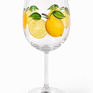 May include: A clear wine glass decorated with hand-painted lemons and green leaves. The yellow lemons are whole and sliced, with brown stems. The design is repeated around the glass, creating a summery, citrus theme.