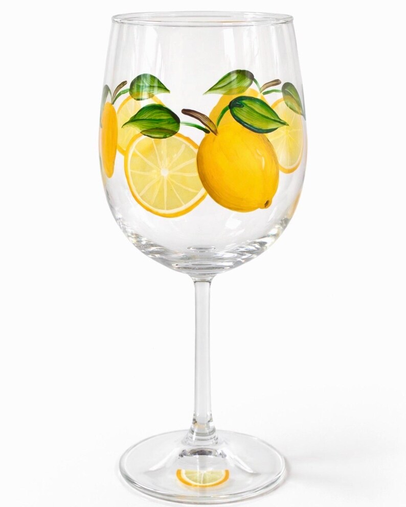 May include: A clear wine glass decorated with hand-painted lemons and green leaves. The yellow lemons are whole and sliced, with brown stems. The design is repeated around the glass, creating a summery, citrus theme.
