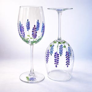 May include: Two clear wine glasses with hand-painted lavender sprigs in shades of purple and blue. The stems and bases are clear. One glass is upright, the other is inverted, showcasing the floral design.