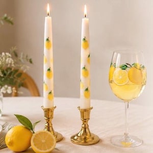May include: Two lit white candles decorated with yellow lemons, set in gold candle holders, next to a wine glass with a lemon design and a lemon slice. A whole lemon and a half lemon are on the table.