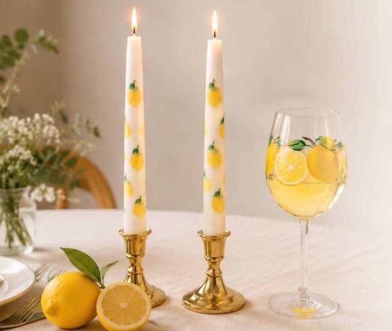 May include: Two lit white candles decorated with yellow lemons, set in gold candle holders, next to a wine glass with a lemon design and a lemon slice. A whole lemon and a half lemon are on the table.