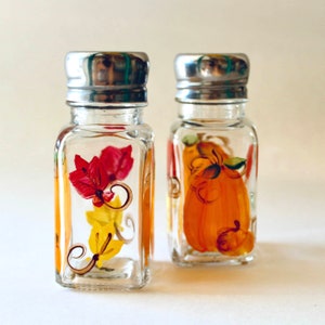 May include: Two clear glass salt and pepper shakers with silver lids. The shakers are hand-painted with fall-themed designs. One shaker features a pumpkin and the other features leaves.