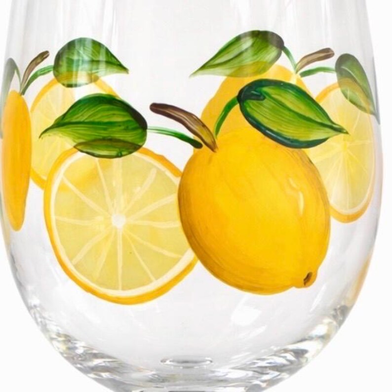 May include: A clear wine glass decorated with hand-painted lemons and green leaves. The design features whole lemons and lemon slices in shades of yellow and green, creating a fresh, summery look.