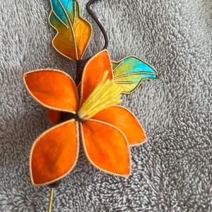 May include: A handmade orange flower with yellow center and green leaves, attached to a brown wire stem with a gold pin.