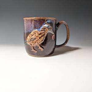 May include: A dark brown ceramic mug featuring a raised, textured relief of a roadrunner bird. The mug has a glossy, mottled glaze with blue and purple undertones, a curved handle, and a wide rim. The bird is depicted in shades of tan and brown.