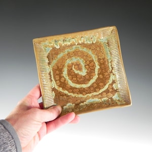 May include: A square ceramic plate with a brown and green glaze. The glaze has a swirling pattern in the center of the plate.