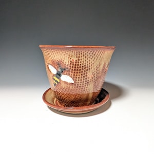 May include: A ceramic plant pot with a matching saucer, featuring a textured honeycomb pattern in shades of brown and tan. A painted bee with yellow and black stripes and white wings is visible on the exterior surface of the tapered vessel.