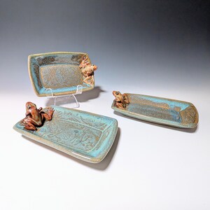 May include: Three ceramic trays with a frog design. The trays are rectangular with a turquoise and brown glaze. Each tray has a raised frog figure on one end. The trays have a floral pattern on the inside.