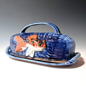 May include: A ceramic butter dish with a lid and handle. The dish is glazed in a deep blue color, with a raised relief of a goldfish in orange, black, and white. The base is a light brown color.