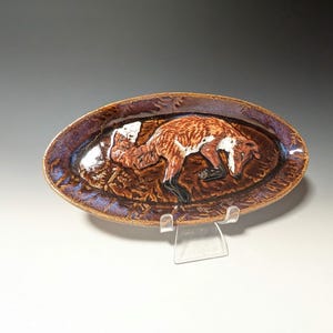 May include: Oval ceramic serving dish with a raised relief of a fox. The fox is brown, white, and black, lying on a textured brown surface. The dish has a dark purple rim with blue accents. The dish is displayed on a clear plastic stand.
