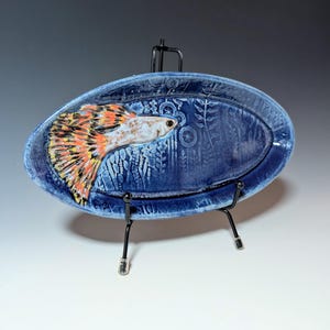 May include: A handcrafted, oval-shaped ceramic dish in a deep blue hue, featuring a detailed relief of a colorful fish with orange, yellow, and black accents. The dish is supported by a black metal stand, showcasing the aquatic-themed art piece.