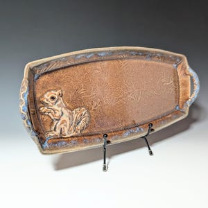 May include: Brown ceramic serving tray with a raised squirrel design. The rectangular tray has a textured surface with a blue rim and handles. The squirrel is in a sitting position, holding something in its paws.