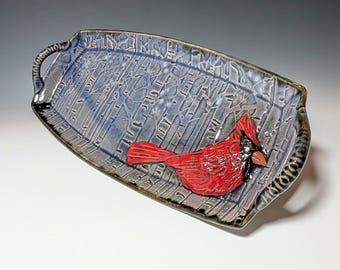 Puede incluir: Una bandeja de cerámica rectangular con una superficie azul texturizada y un pájaro cardenal rojo en relieve. La bandeja tiene asas y mide aproximadamente 30 cm de largo.