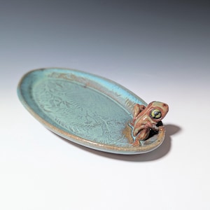 May include: An oval ceramic dish with a teal glaze and a brown rim. The dish features a detailed embossed pattern and a small, brown frog figurine on the edge. The frog has yellow eyes and is perched on the rim.