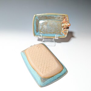 May include: Two handmade ceramic rectangular dishes. One dish is upright, showing a frog figurine on the side. The other dish is upside down, revealing a textured pattern. Both have a turquoise rim and a brown interior.