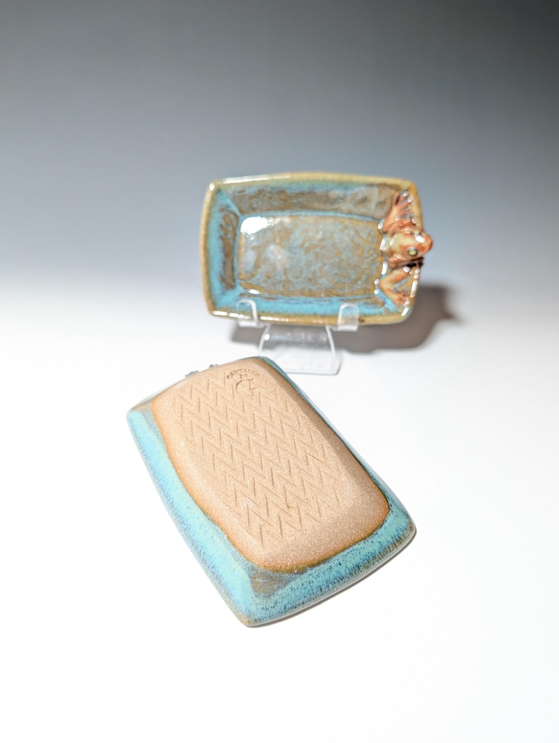 May include: Two handmade ceramic rectangular dishes. One dish is upright, showing a frog figurine on the side. The other dish is upside down, revealing a textured pattern. Both have a turquoise rim and a brown interior.