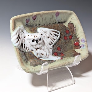 May include: A rectangular ceramic tray with a light green glazed border and a textured brown center. A raised white owl with spread wings is attached to the left side, while small red circular accents are scattered across the brown surface.