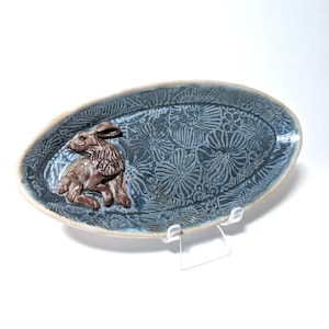 May include: Oval ceramic serving dish with a blue floral pattern and a brown rabbit figurine. The dish has a light blue rim and is displayed on a clear stand. A decorative piece for the home.