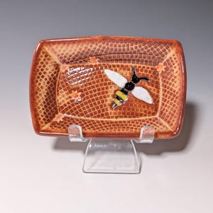Puede incluir: Un plato rectangular de cer&aacute;mica con una superficie marr&oacute;n texturizada y un dise&ntilde;o de abeja. La abeja tiene alas blancas, cuerpo negro y rayas amarillas. El plato est&aacute; sostenido por un soporte de pl&aacute;stico transparente.