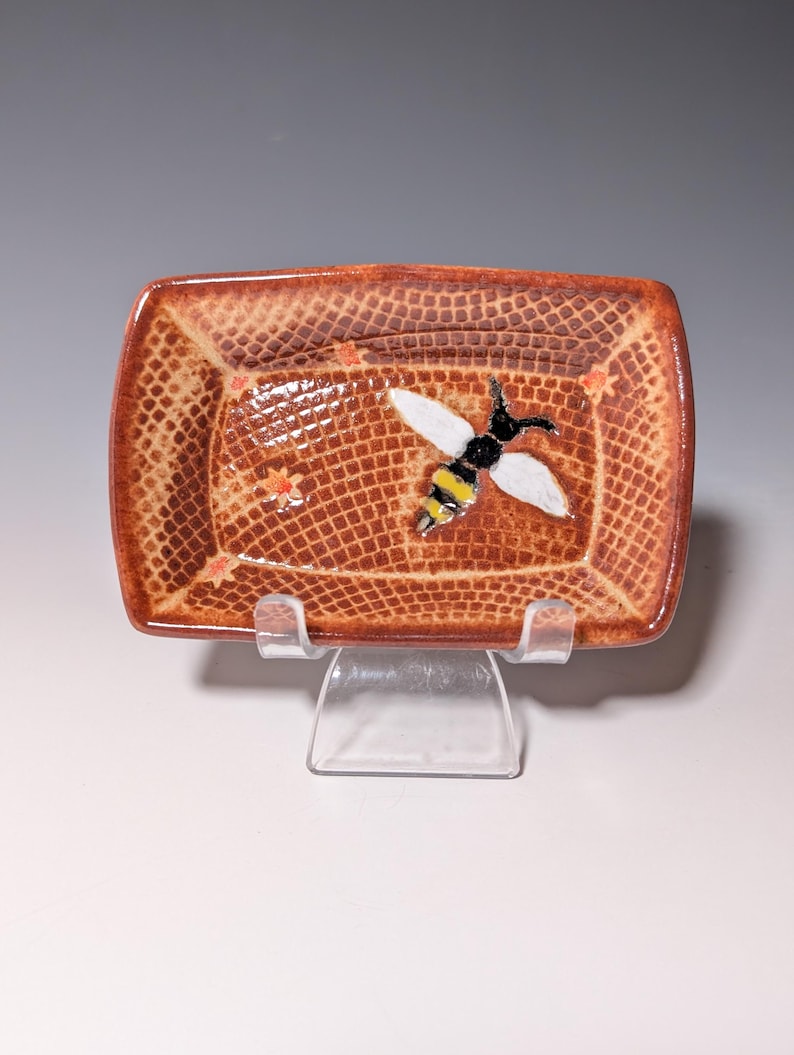 Puede incluir: Un plato rectangular de cer&aacute;mica con una superficie marr&oacute;n texturizada y un dise&ntilde;o de abeja. La abeja tiene alas blancas, cuerpo negro y rayas amarillas. El plato est&aacute; sostenido por un soporte de pl&aacute;stico transparente.