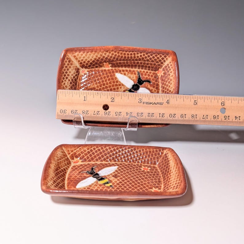 Puede incluir: Dos platos rectangulares de cer&aacute;mica con un patr&oacute;n marr&oacute;n texturizado e ilustraciones de abejas. Los platos miden aproximadamente 10 cm de largo. Un plato est&aacute; colocado encima del otro, con una regla colocada sobre ellos.