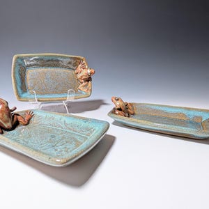 May include: Three ceramic trays with frog figurines. The trays are rectangular and oval, with a turquoise and brown glaze. Each tray features a detailed frog sculpture on the edge. The frog sculptures are brown and add a whimsical touch to the functional art.