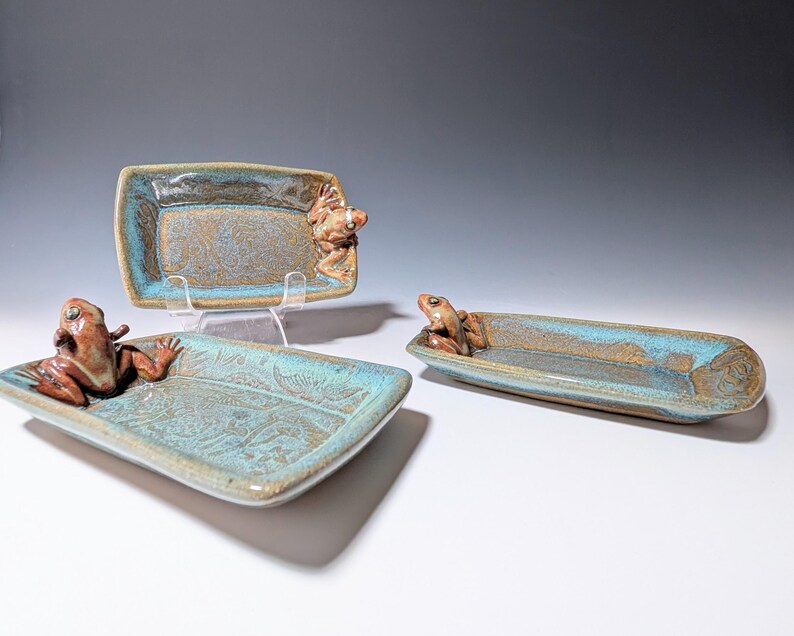 May include: Three ceramic trays with frog figurines. The trays are rectangular and oval, with a turquoise and brown glaze. Each tray features a detailed frog sculpture on the edge. The frog sculptures are brown and add a whimsical touch to the functional art.