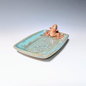 May include: A rectangular ceramic dish with a turquoise rim and a brown frog figurine. The dish has a textured surface with a detailed pattern. The frog is brown and appears to be resting on the edge of the dish.