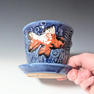 May include: A blue ceramic planter with a matching saucer. The planter features a raised relief design of a goldfish in shades of orange, white, and black. The planter has a textured surface and is suitable for small plants.