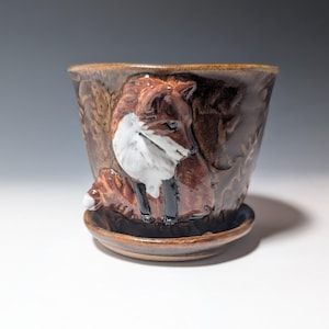 May include: A brown ceramic planter with a glossy finish featuring a raised, sculpted fox figure on the front. The fox has a white chest and face with dark brown and black details. The planter includes a matching circular saucer base.