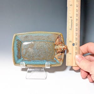 May include: A rectangular ceramic dish with a frog figurine. The dish has a brown and teal glaze with a textured surface. The frog is brown with green eyes. The dish measures approximately 6 inches long.