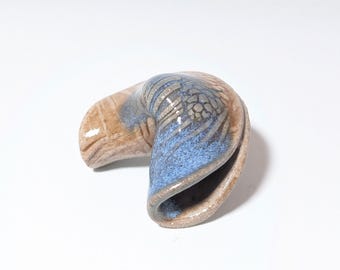Puede incluir: Una escultura de cerámica con forma de galleta de la fortuna. La galleta presenta una mezcla de colores beige, azul y gris. La sección azul tiene un aspecto texturizado, mientras que la sección beige tiene un acabado liso. La escultura está sobre un fondo blanco.