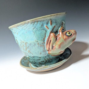 May include: A ceramic planter with a turquoise glaze and a frog sculpture attached. The frog is brown and green with yellow eyes. The planter has a matching saucer.