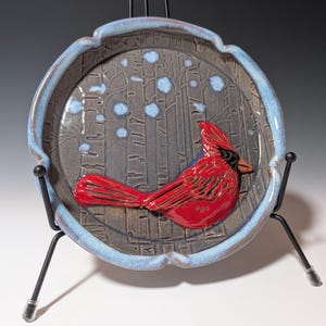 May include: A ceramic plate with a raised red cardinal design. The plate has a blue rim and a dark gray center with a tree and snow design. The plate is displayed on a black metal stand.