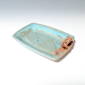 May include: A rectangular ceramic dish with a turquoise glaze and a brown frog figurine on the edge. The dish has a textured surface with a leaf pattern. The frog is brown with green eyes.