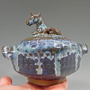 May include: A handmade ceramic jar with a lid, featuring a blue and brown glaze. The lid is topped with a small, detailed unicorn figurine. The jar has a round shape with handles and a textured surface, showcasing a unique, artistic design.