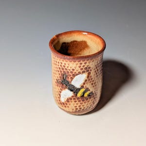 May include: A small, handmade ceramic cup with a bee design. The cup has a textured, honeycomb-like pattern in shades of tan and brown. A bee with white wings and yellow stripes is painted on the front. The rim is a darker brown.