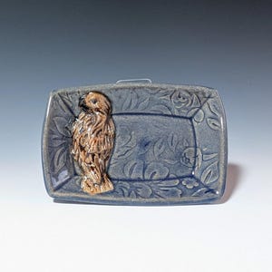 May include: A small, rectangular ceramic dish with a blue glaze and a raised, brown hawk design. The dish has a floral pattern.