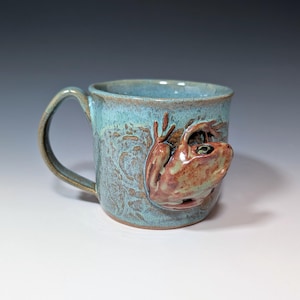 May include: A handmade ceramic mug with a teal glaze and a sculpted frog design. The frog is a reddish-brown color and appears to be climbing the side of the mug. The mug has a handle and a textured surface.