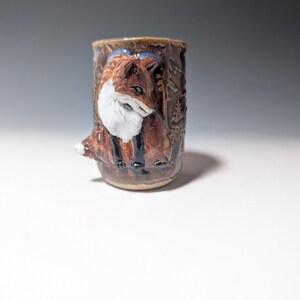 May include: A cylindrical ceramic mug featuring a raised, three-dimensional relief of a fox with reddish-brown fur, a white chest, and black legs. The mug has a glossy brown glaze and depicts a forest scene with trees embossed around the fox.